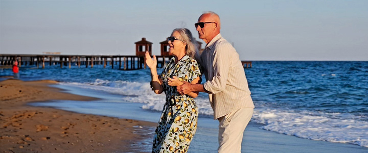 Ein älteres Pärchen mit Sonnenbrillen steht am Strand und blickt für ein Foto nach links, während im Hintergrund das Meer und ein Steg zu sehen sind.